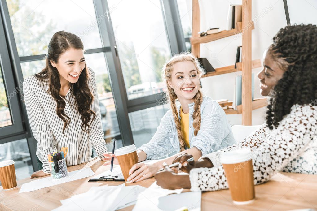 multicultural cheerful businesswomen with coffee to go having conversation in office