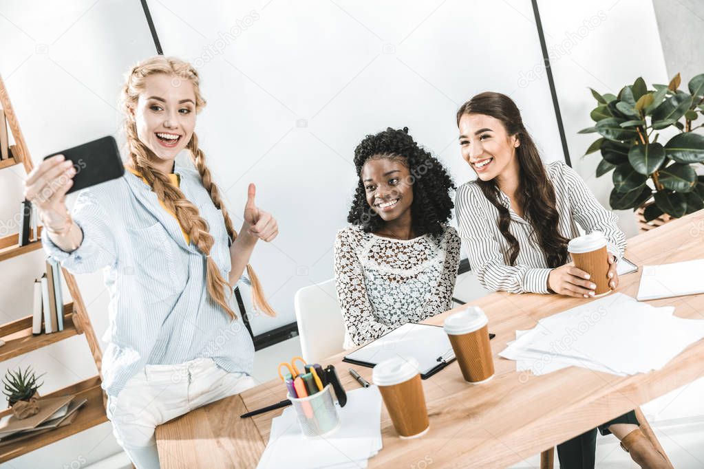 multicultural smiling businesswomen taking selfie on smartphone at workplace