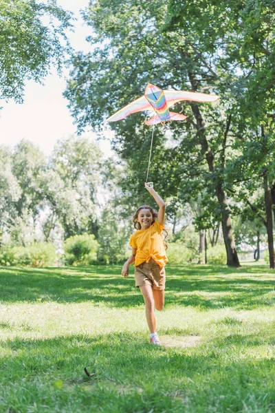 カラフルな凧を押しながら公園で芝生の上を実行しているカメラで笑顔の美しい幸せな子供 — ストック写真