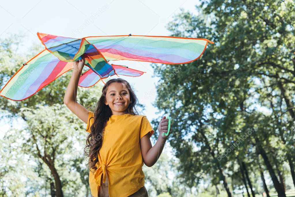 low angle view of happy child holding colorful kite and smiling at camera in park