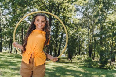beautiful happy kid holding hula hoop and smiling at camera in park  clipart