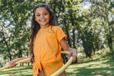adorable happy child playing with hula hoop and smiling at camera in park clipart
