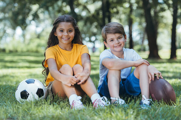 cute happy children with soccer and rugby balls sitting and smiling at camera in park