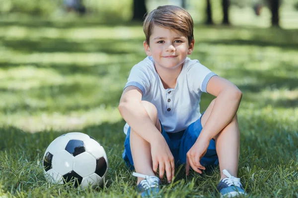 サッカー ボールと草の上に座って 公園にカメラで笑顔のかわいい男の子 — ストック写真