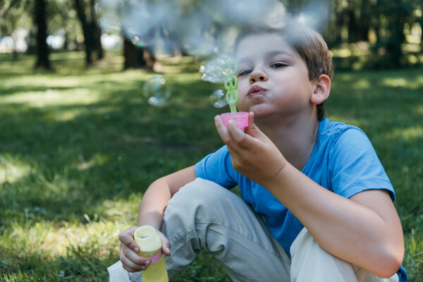 adorable child blowing soap bubbles while sitting on grass in park