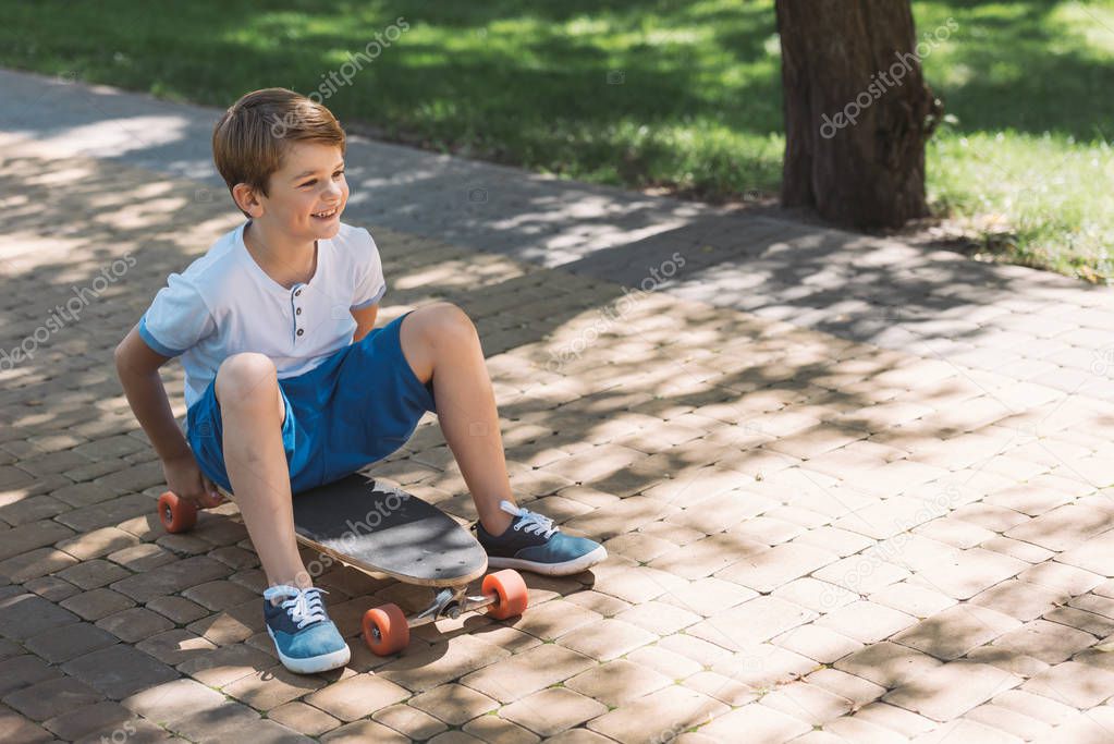 high angle view of smiling boy sitting on skateboard and looking away in park
