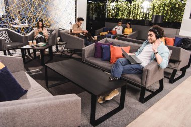dizüstü bilgisayar ile kanepede oturan ve el sallayarak modern coworking ofiste Kulaklığımda genç işadamı 