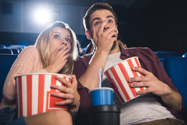 scared couple with popcorn and soda drink watching film together in cinema