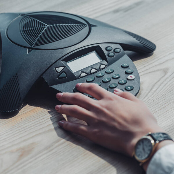 cropped shot of businesswoman pushing button of speakerphone on wooden table at office