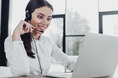 beautiful female call center worker with headphones and laptop sitting at workplace clipart