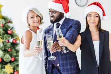 selective focus of multiethnic businesspeople in santa hats clinking with glasses of champagne in office