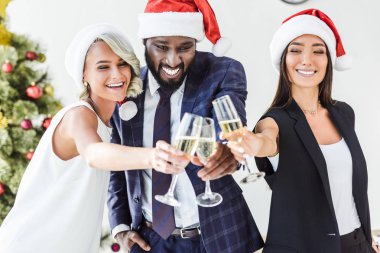 smiling multicultural businesspeople in santa hats clinking with glasses of champagne in office