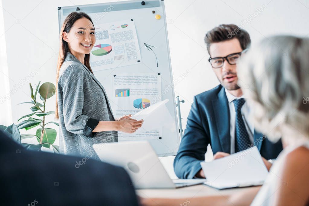 smiling asian businesswoman standing near flipchart during meeting in office