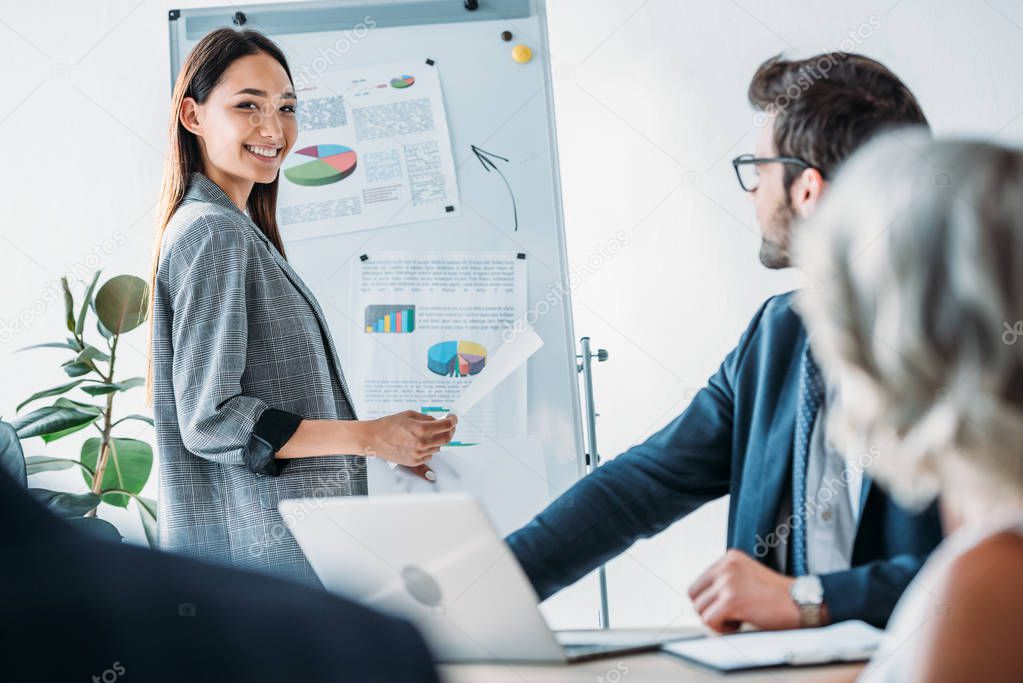 smiling asian businesswoman standing near flipchart during meeting in office and looking at camera