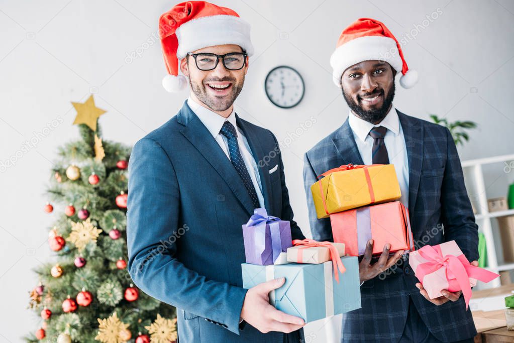 smiling multicultural businessmen in santa hats holding gift boxes in office