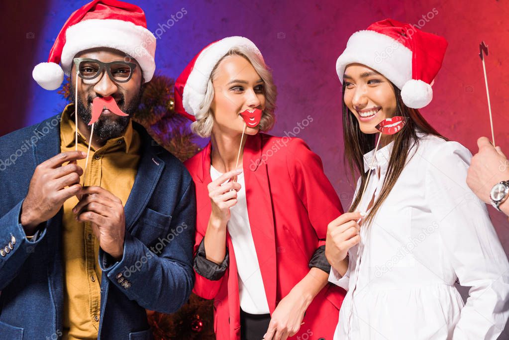 happy multicultural businesspeople holding lips and glasses on sticks at new year corporate party