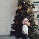 Cute kid in santa hat standing near christmas tree at home