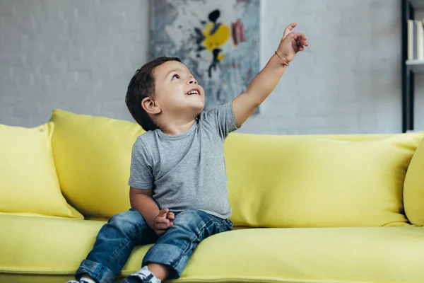 微笑的小男孩在家里坐在黄色的沙发上指着 — 图库照片