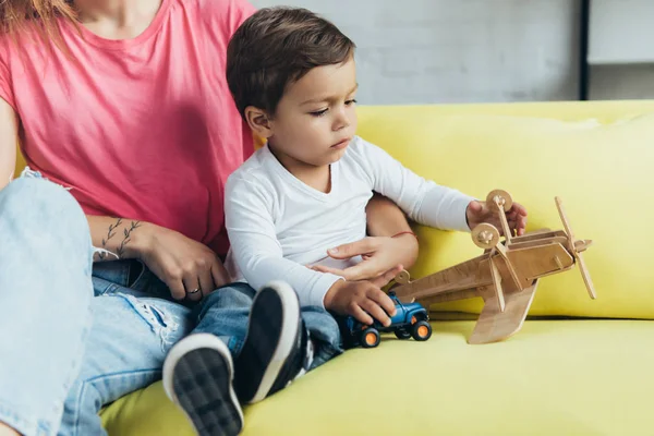 Little Son Playing Toy Car Wooden Plane While Sitting Sofa — Free Stock Photo