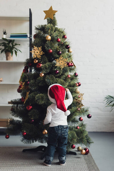 вид сзади на ребенка в шляпе Санты, украшающего елку дома

