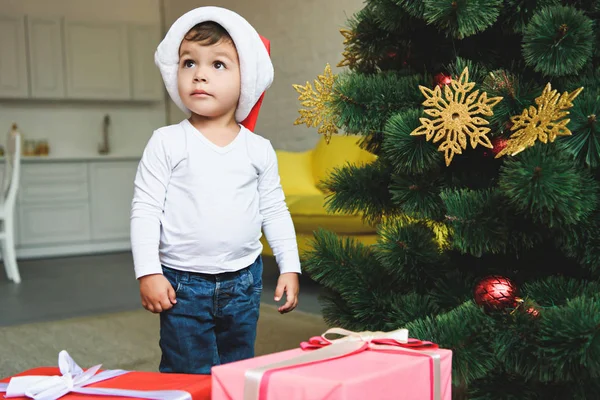 ギフト用の箱のクリスマス ツリーのそばでサンタの帽子で愛らしい小さな子供 — ストック写真