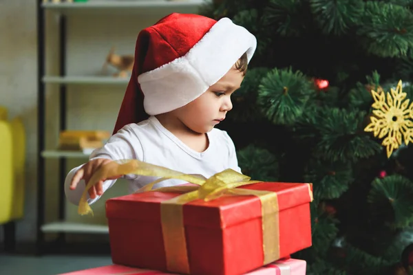 ギフト クリスマス ツリーのそばでサンタの帽子で愛らしい幼児  — 無料ストックフォト