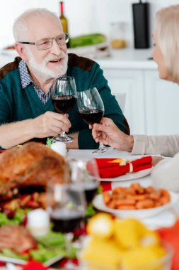 smiling senior couple clinking by wine glasses at serve table with baked turkey on thanksgiving  clipart