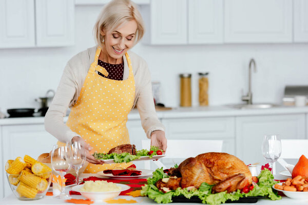 smiling senior woman carrying meal on table with delicious dishes for thanksgiving celebration 