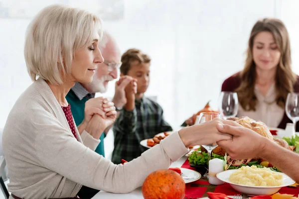 selective focus of grandmother praying with family at served table with turkey before holiday dinner on thanksgiving