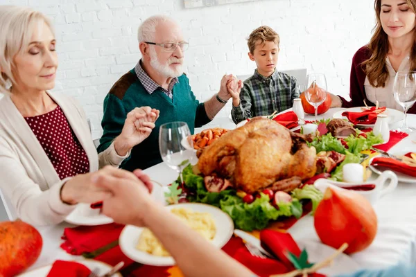 happy family holding hands and praying at served table with turkey before holiday dinner on thanksgiving