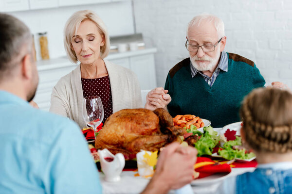 senior couple with family praying at served table with turkey before holiday dinner on thanksgiving 