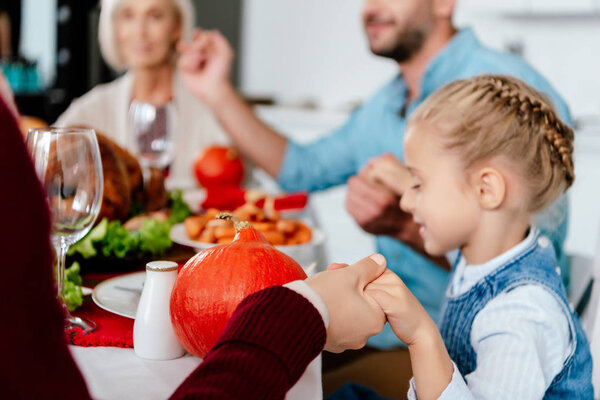 selective focus of adorable kid holding hands and praying with family at served table on thanksgiving 