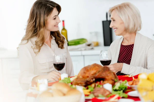 happy adult woman with wine glass talking to senior mother at served table with baked turkey on thanksgiving