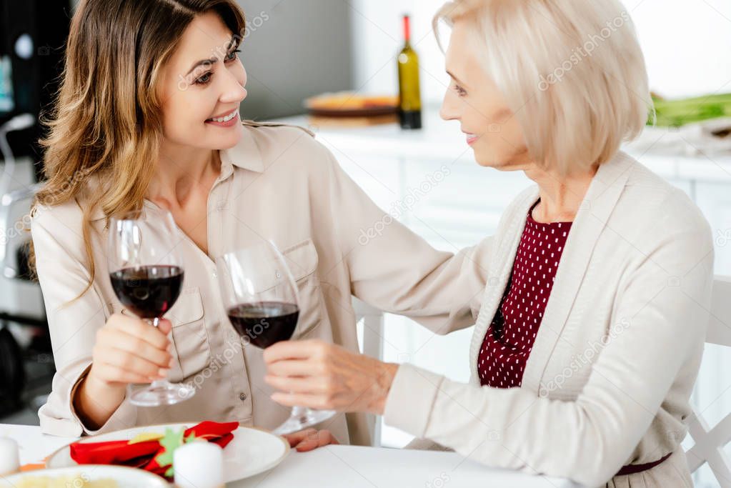 smiling adult woman clinking by wine glasses with senior mother at served table for thanksgiving celebration 