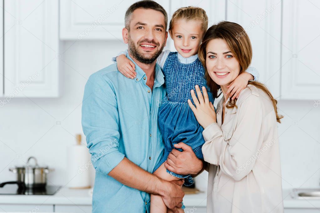 portrait of happy family holding and embracing little daughter and looking at camera at kitchen 