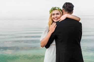 çelenk ve damat takım elbise kumsalda sarılma içinde çekici gülümseyen gelin