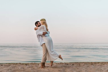 bride in white dress and groom cuddling on beach clipart