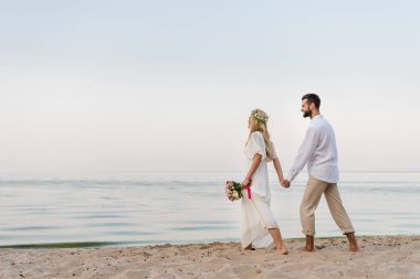 Gelin ve damat elele ve düğün buket ile sahilde yürüyüş yan görünüm
