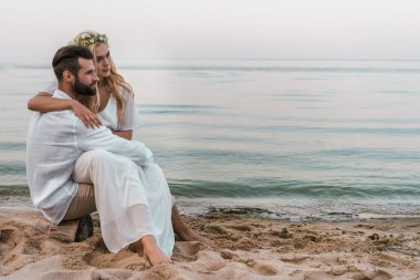 elegant groom and bride sitting on log on beach and looking away clipart
