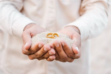 cropped image of groom holding wedding golden rings with sand in hands on beach clipart