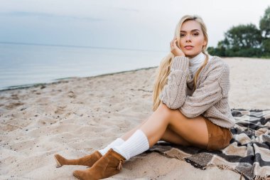 sahilde oturup, çene el üstünde dinlenme ve kameraya bakarak sonbahar kıyafeti çekici sarışın kız