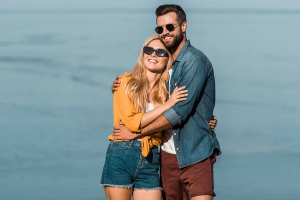 Sonriente Pareja Gafas Sol Abrazándose Pie Cerca Del Océano —  Fotos de Stock