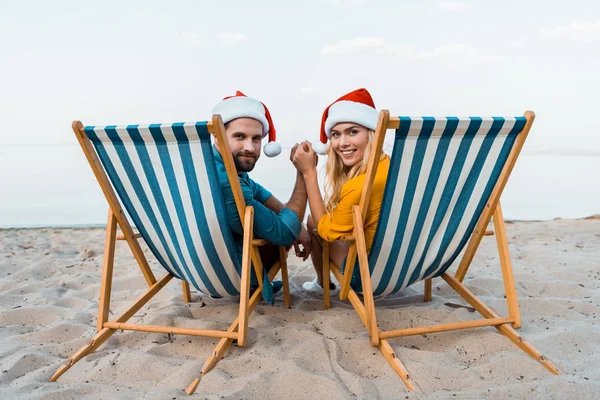 Par Santa Hattar Sitter Solstolar Hålla Händerna Och Titta Kameran — Stockfoto