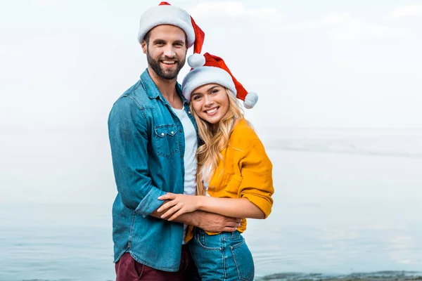 微笑的情侣在圣诞老人的帽子拥抱在海滩上 看着相机 — 图库照片