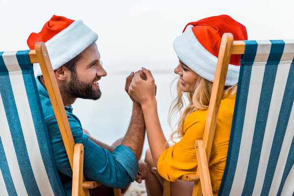 smiling couple in santa hats sitting on sun loungers, holding hands and looking at each other on beach