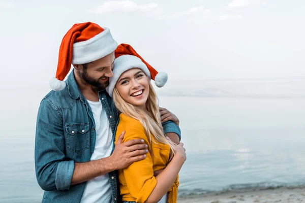 微笑的男朋友在圣诞老人帽子拥抱女朋友在海滩 — 图库照片