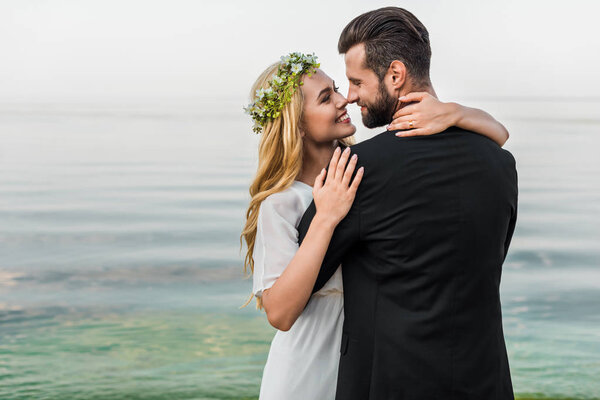 attractive bride in wreath and groom in suit cuddling on beach and touching with noses