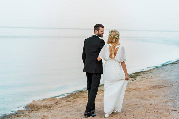 back view of wedding couple holding hands and walking on beach