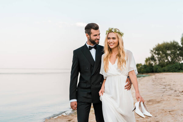 groom hugging bride and walking on beach, bride holding high heels in hand