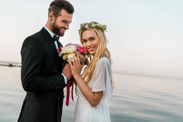 Боковой Вид Свадебную Пару Стоящую Букетом Пляже Улыбающуюся Невесту Смотрящую — стоковое фото